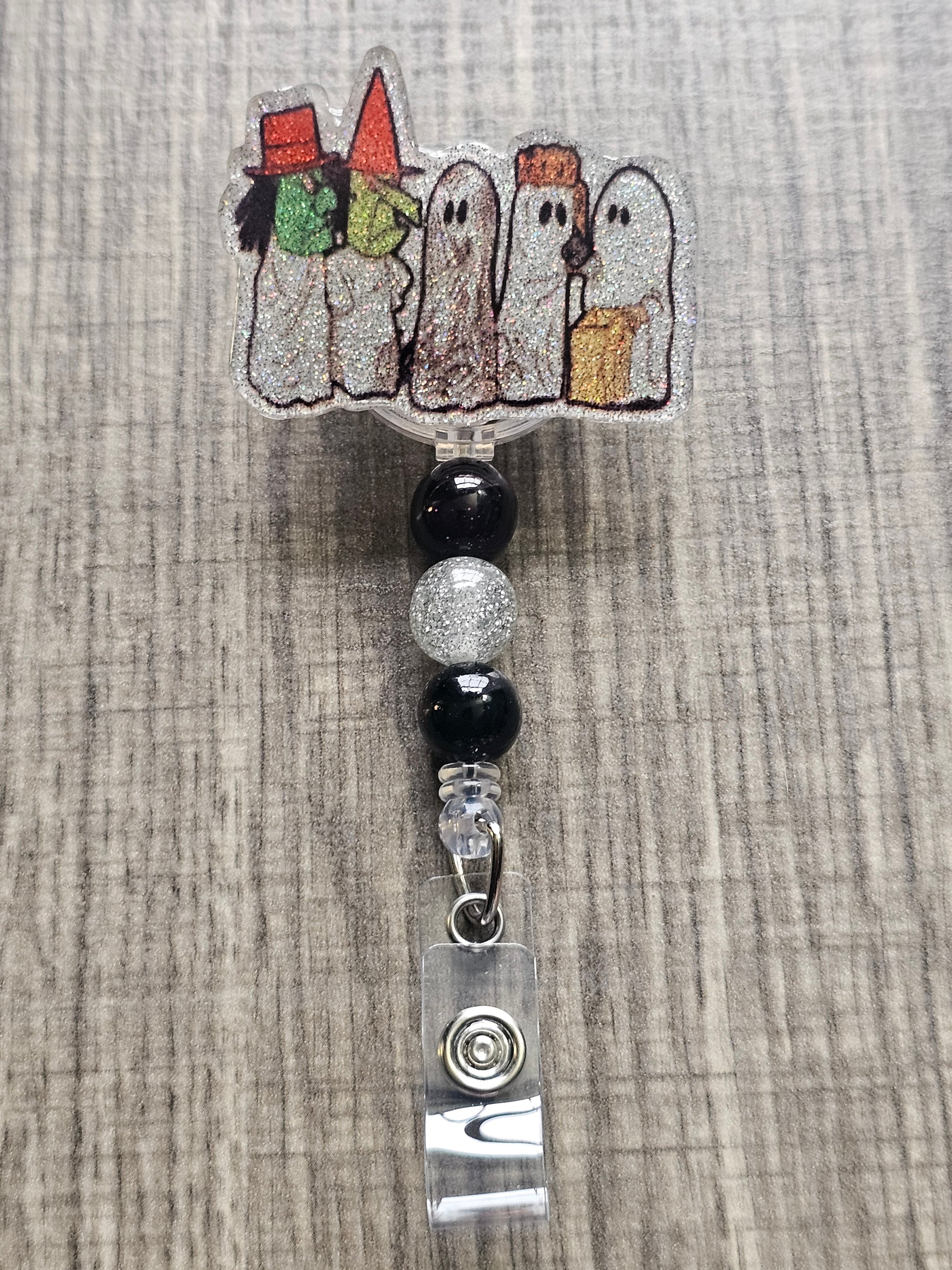 Badge reel depicting five figures in white ghost costumes, two wearing witch masks, and one holding a trick-or-treat bag, with matching decorative beads.