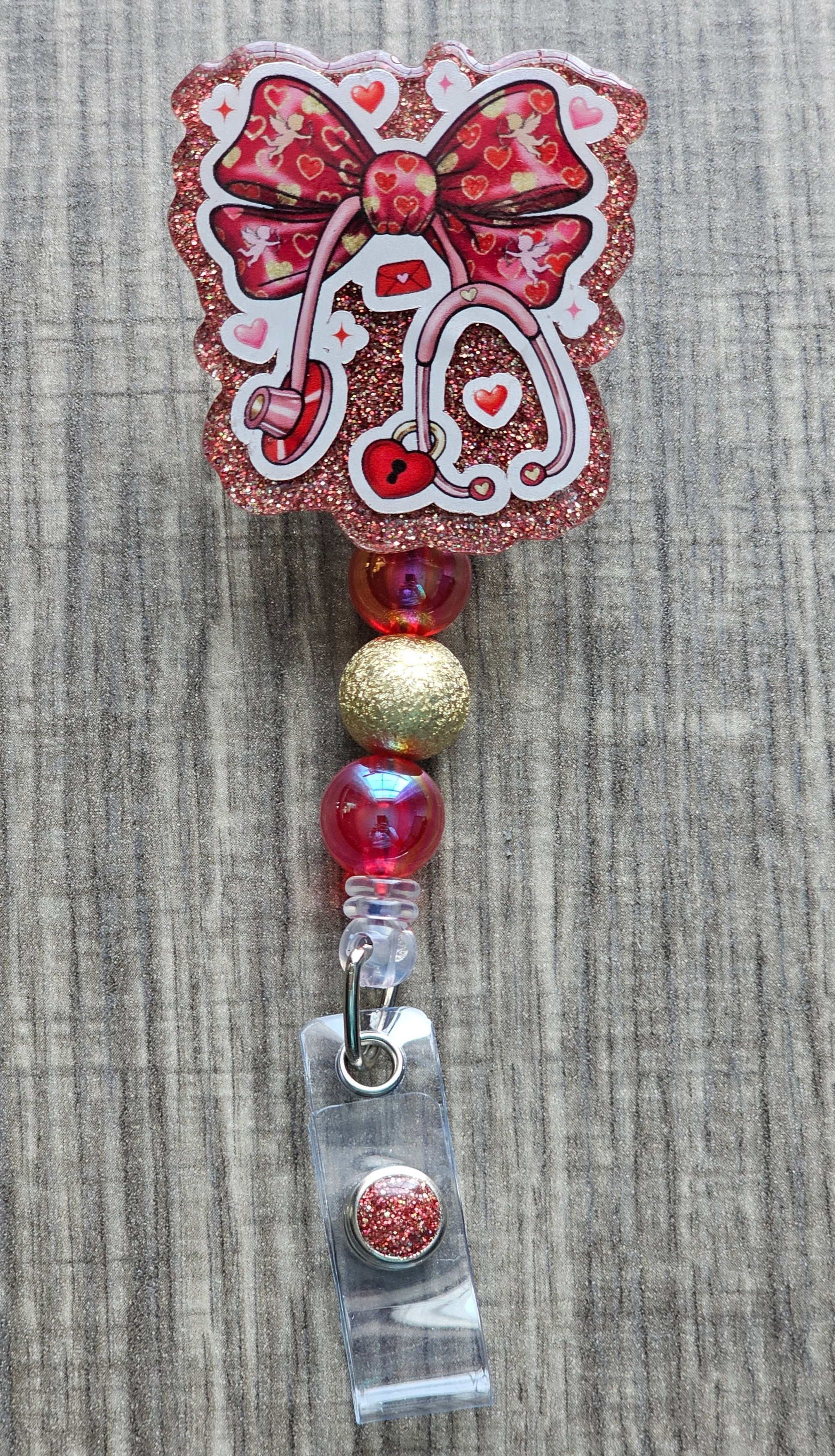 A red and pink glitter retractable nurse badge reel featuring a stethoscope with a heart-shaped lock and bow accent. The badge is embellished with red and gold iridescent beads.