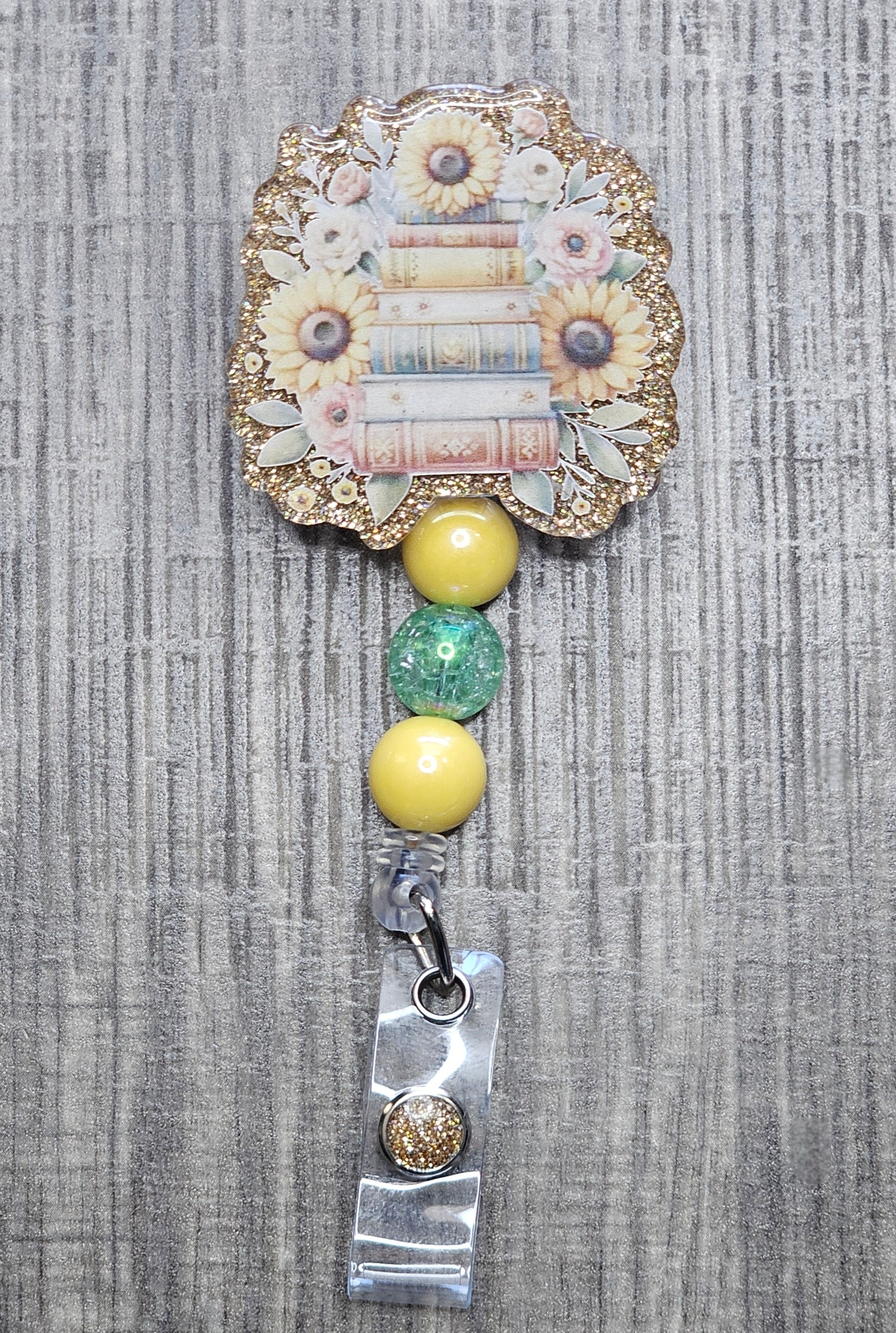 Retractable badge reel showing a stack of books with sunflowers on a gold background with matching yellow beads. 