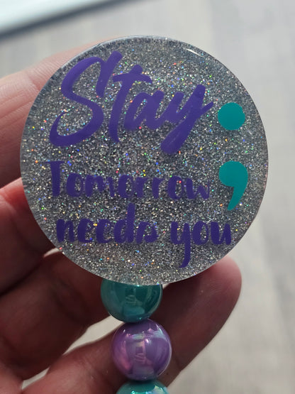 A badge Reel ith a silver background and purple lettering stating stay tomorrow needs you with a turquoise semicolon