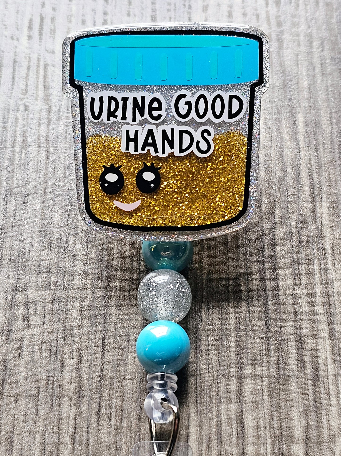 A badge reel featuring a smiling animated urine sample bottle filled with glittery “urine” and the phrase "Urine Good Hands.