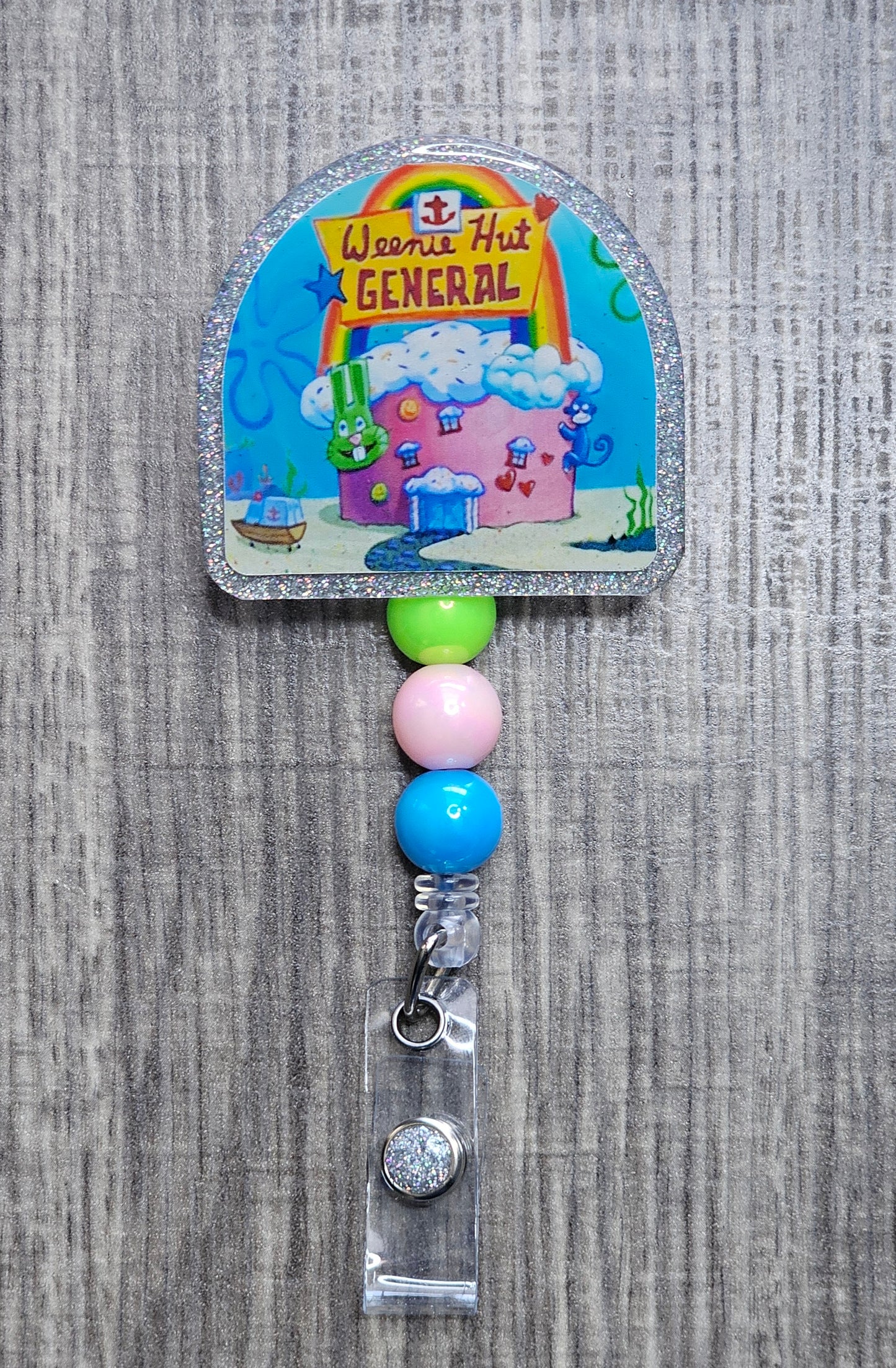 A colorful badge reel featuring the iconic hospital from SpongeBob  'Weenie Hut General'. It includes decorative beads and an alligator clip.
