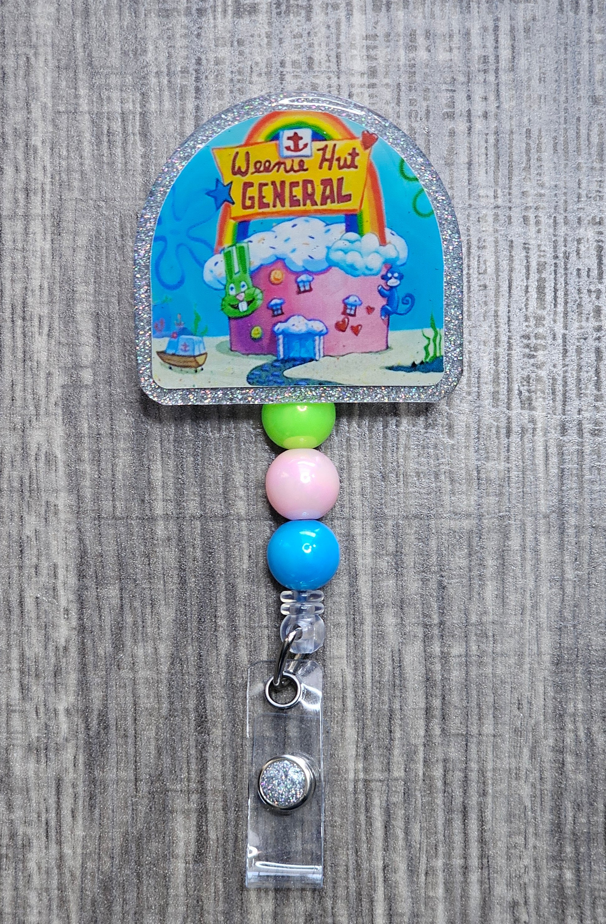 A colorful badge reel featuring the iconic hospital from SpongeBob  'Weenie Hut General'. It includes decorative beads and an alligator clip.