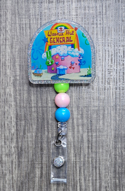 A colorful badge reel featuring the iconic hospital from SpongeBob  'Weenie Hut General'. It includes decorative beads and an alligator clip.