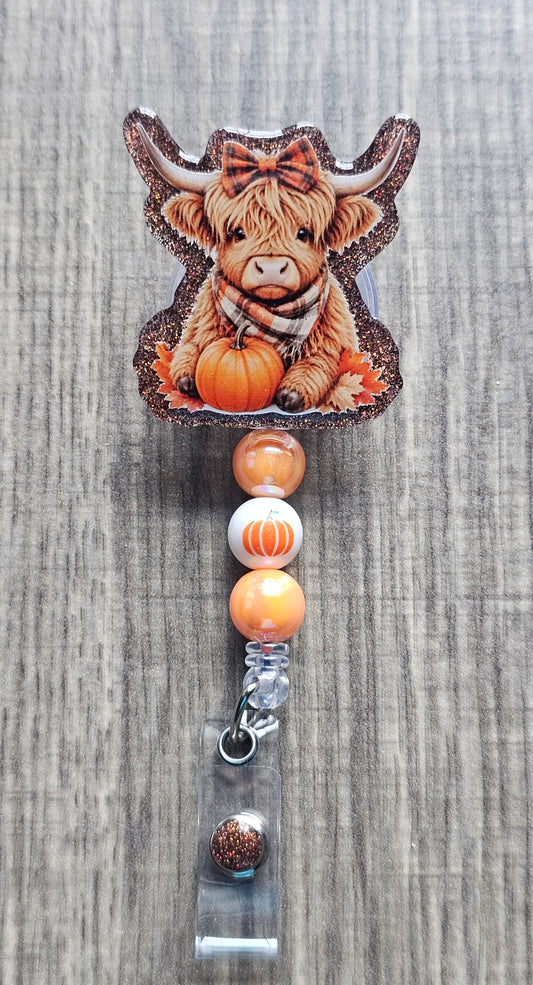  Autumn themed badge reel showcasing a fluffy highland cow with long horns surrounded by pumpkins, fall leaves, and a bow.