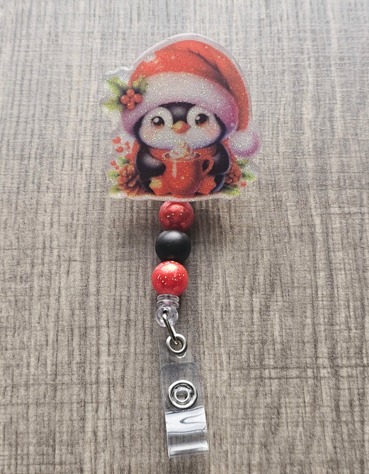 This badge reel features a cheerful penguin, complete with a Christmas hat and a heartwarming smile, holding a cozy cup of hot chocolate.